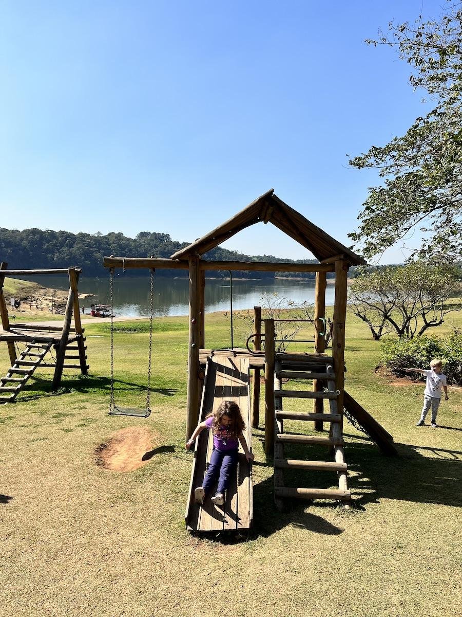 Crianças brincando em playground de madeira ao ar livre, com gramado e represa ao fundo.