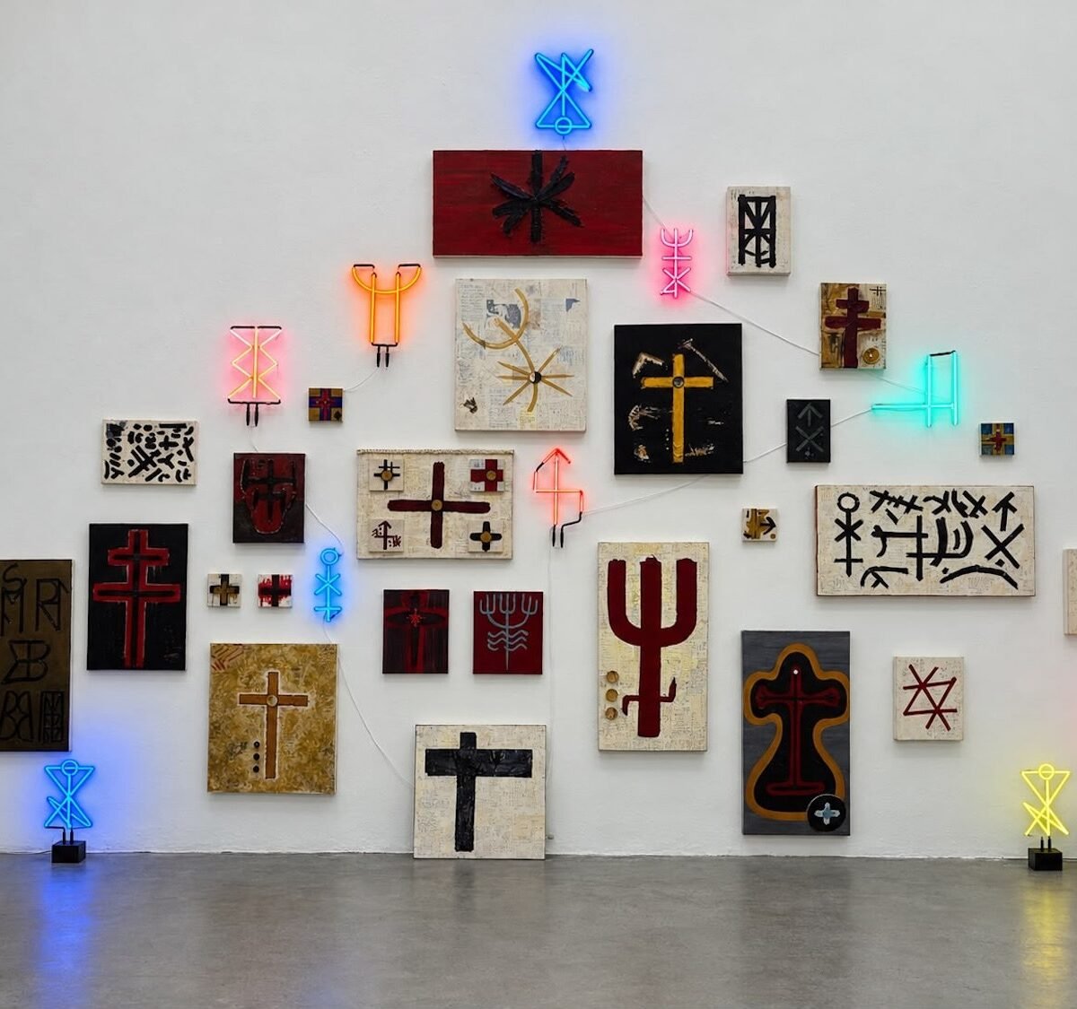 Parede com pinturas, colagens e néons em forma de símbolos de proteção, da exposição 144 de J.P. Cuenca
