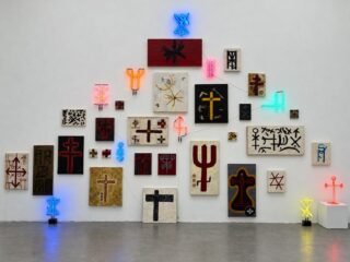 Parede com pinturas, colagens e néons em forma de símbolos de proteção, da exposição 144 de J.P. Cuenca