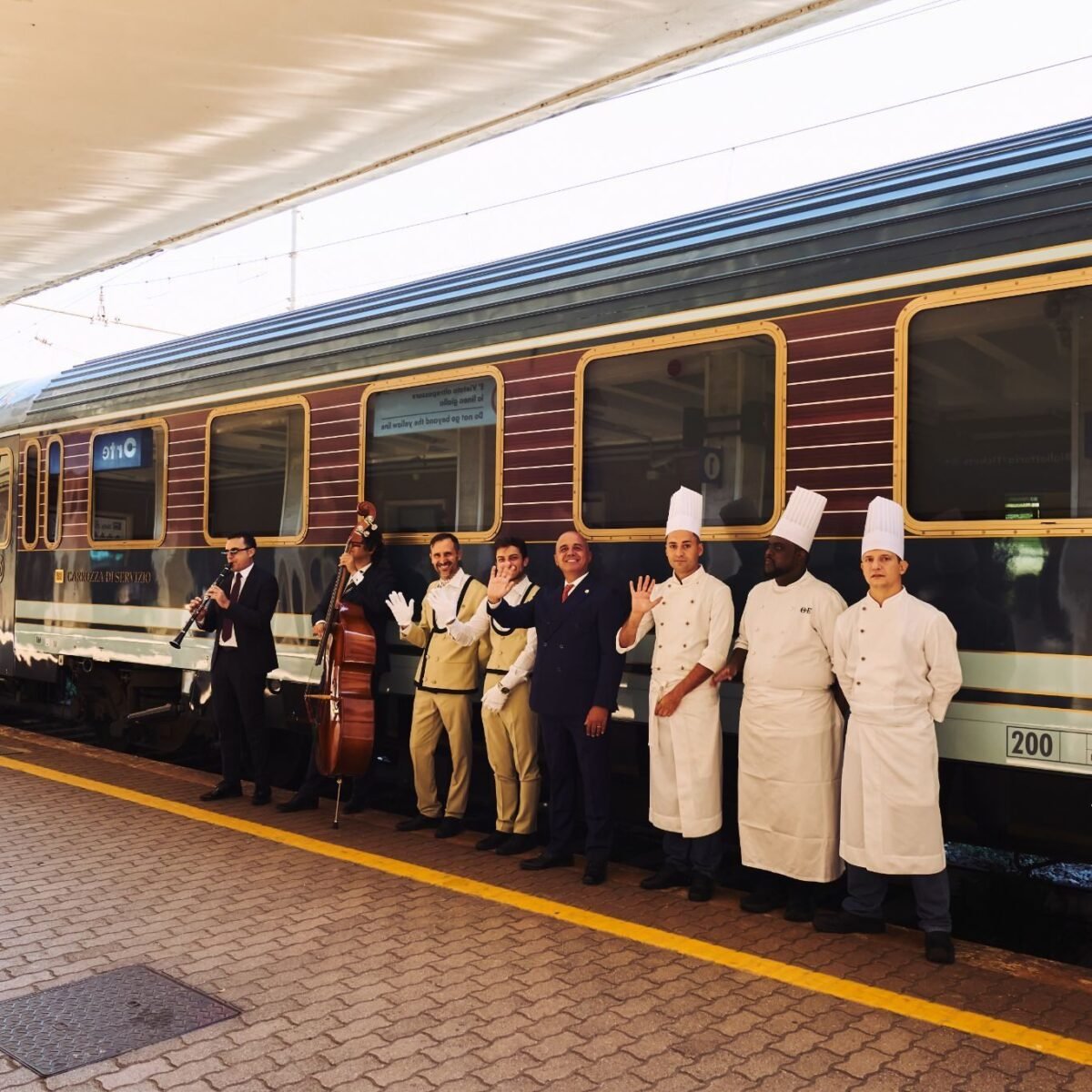 Vagão do La Dolce Vita Orient Express na plataforma, com músicos, equipe de bordo e chefs enfileirados acenando