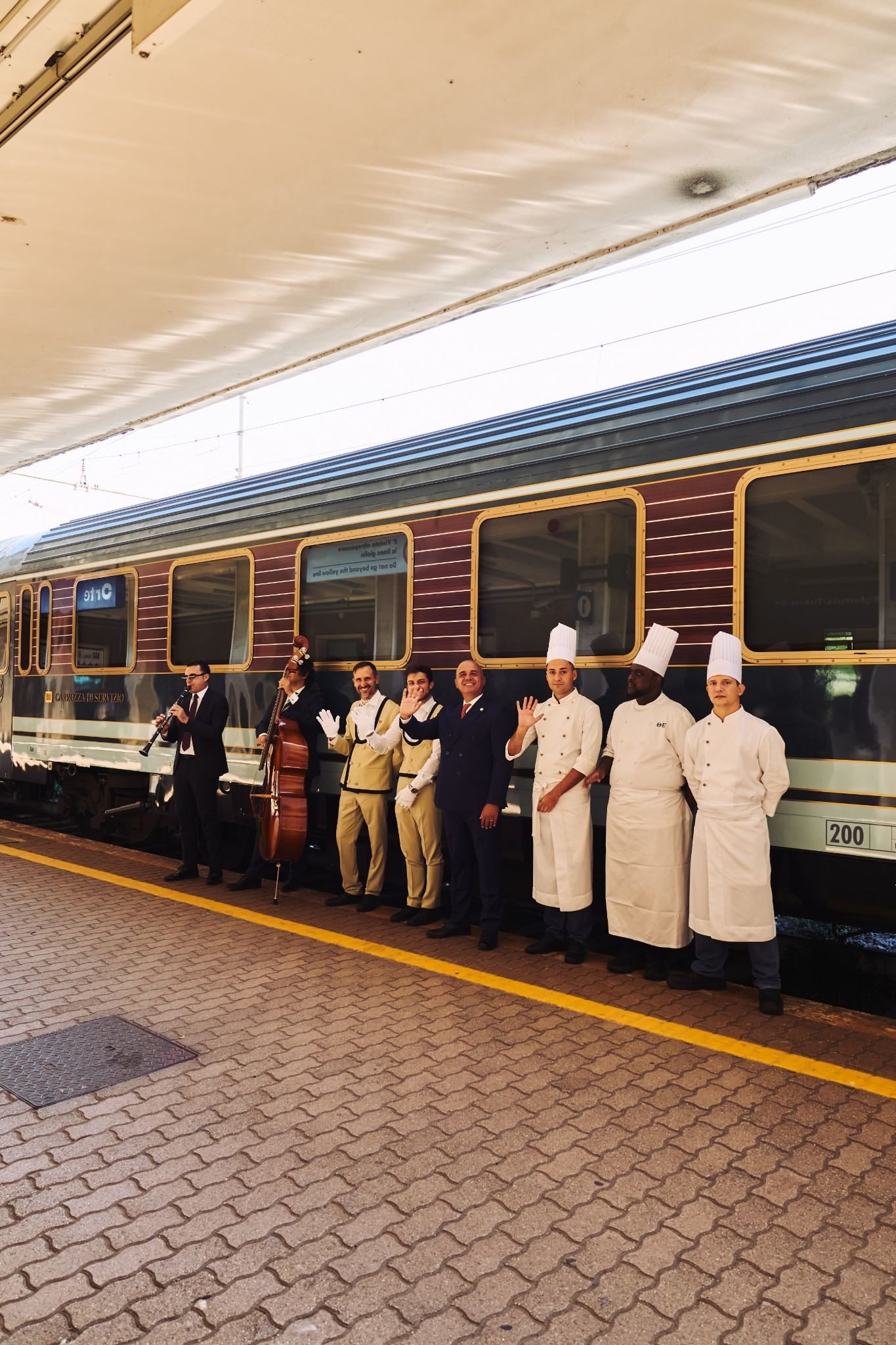 Vagão do La Dolce Vita Orient Express na plataforma, com músicos, equipe de bordo e chefs enfileirados acenando