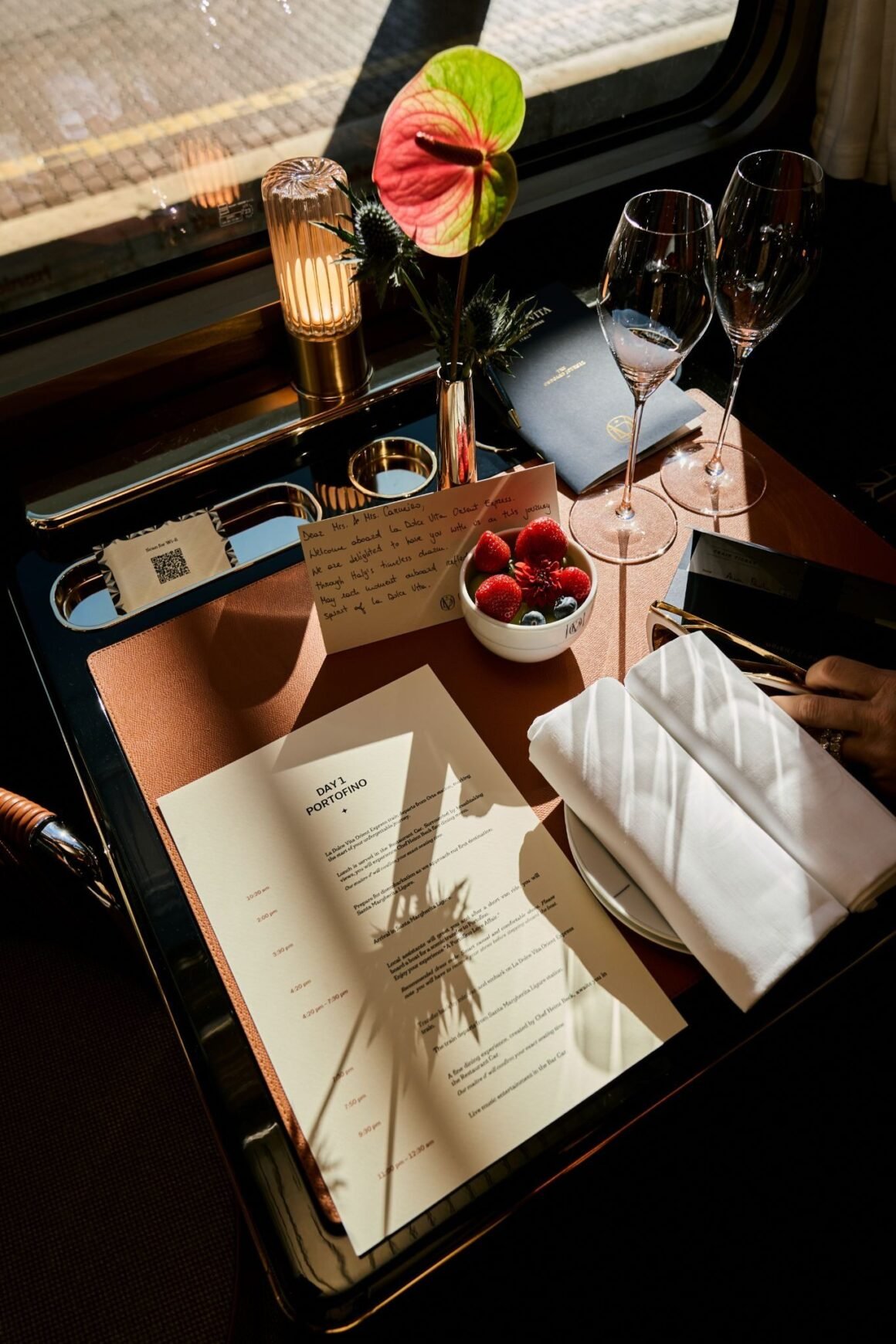 Mesa a bordo do La Dolce Vita Orient Express com taças, cartão de boas-vindas, frutas vermelhas e menu do dia 1, Portofino