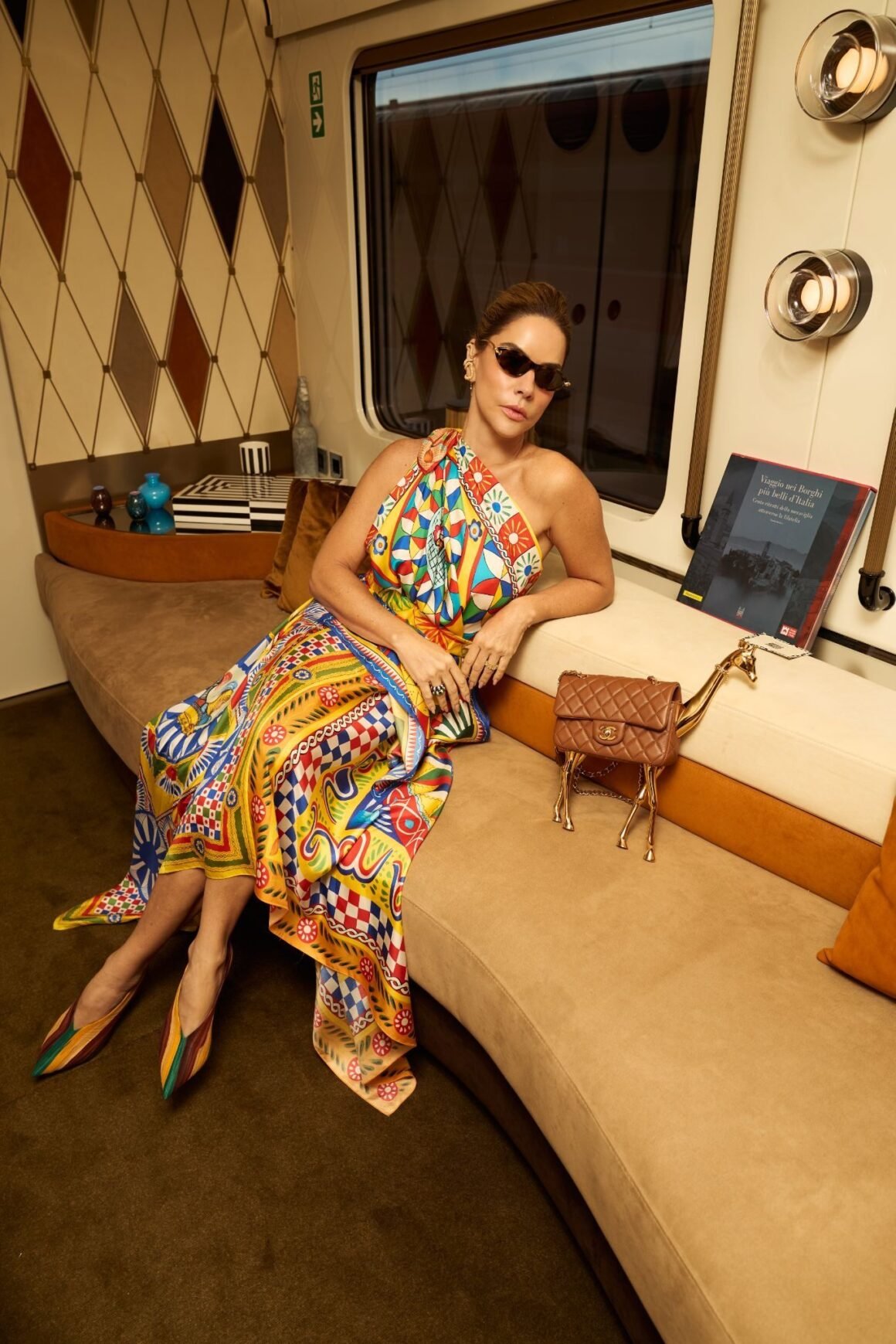 Ana Paula Carneiro sentada em sofá de cabine do La Dolce Vita Orient Express, com lenço estampado e bolsa marrom