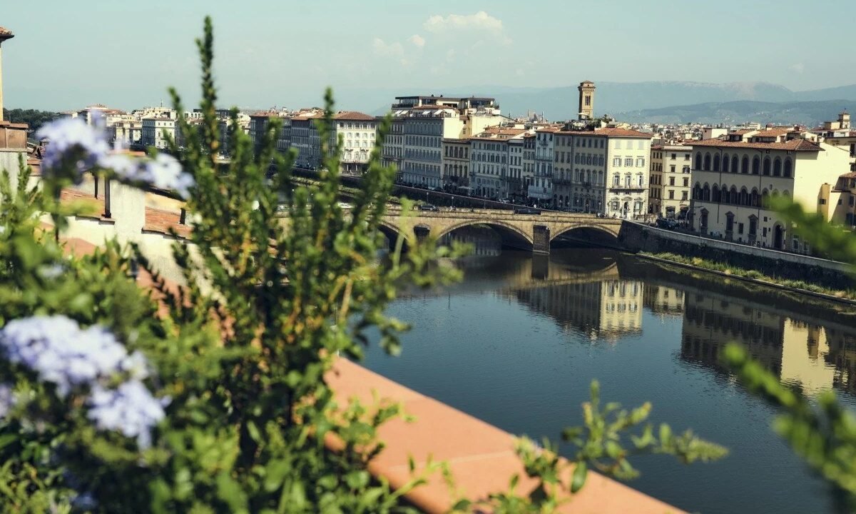 Vista do rio Arno e de uma ponte de pedra em Florença, com flores e folhagem em primeiro plano