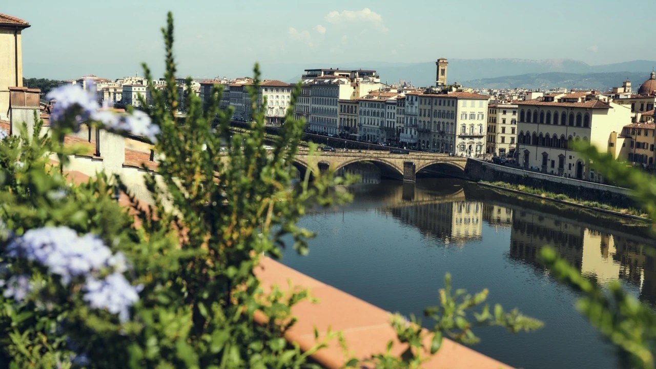 Vista do rio Arno e de uma ponte de pedra em Florença, com flores e folhagem em primeiro plano