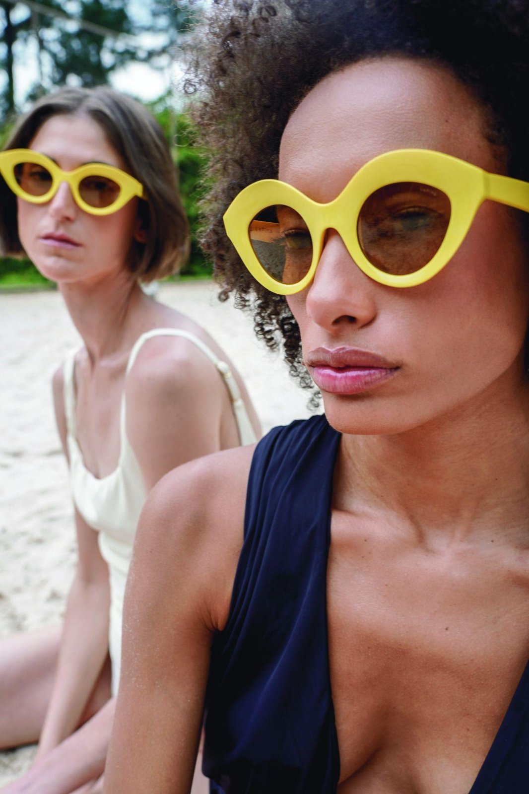 Two women wearing yellow sculptural LAPIMA sunglasses on a beach