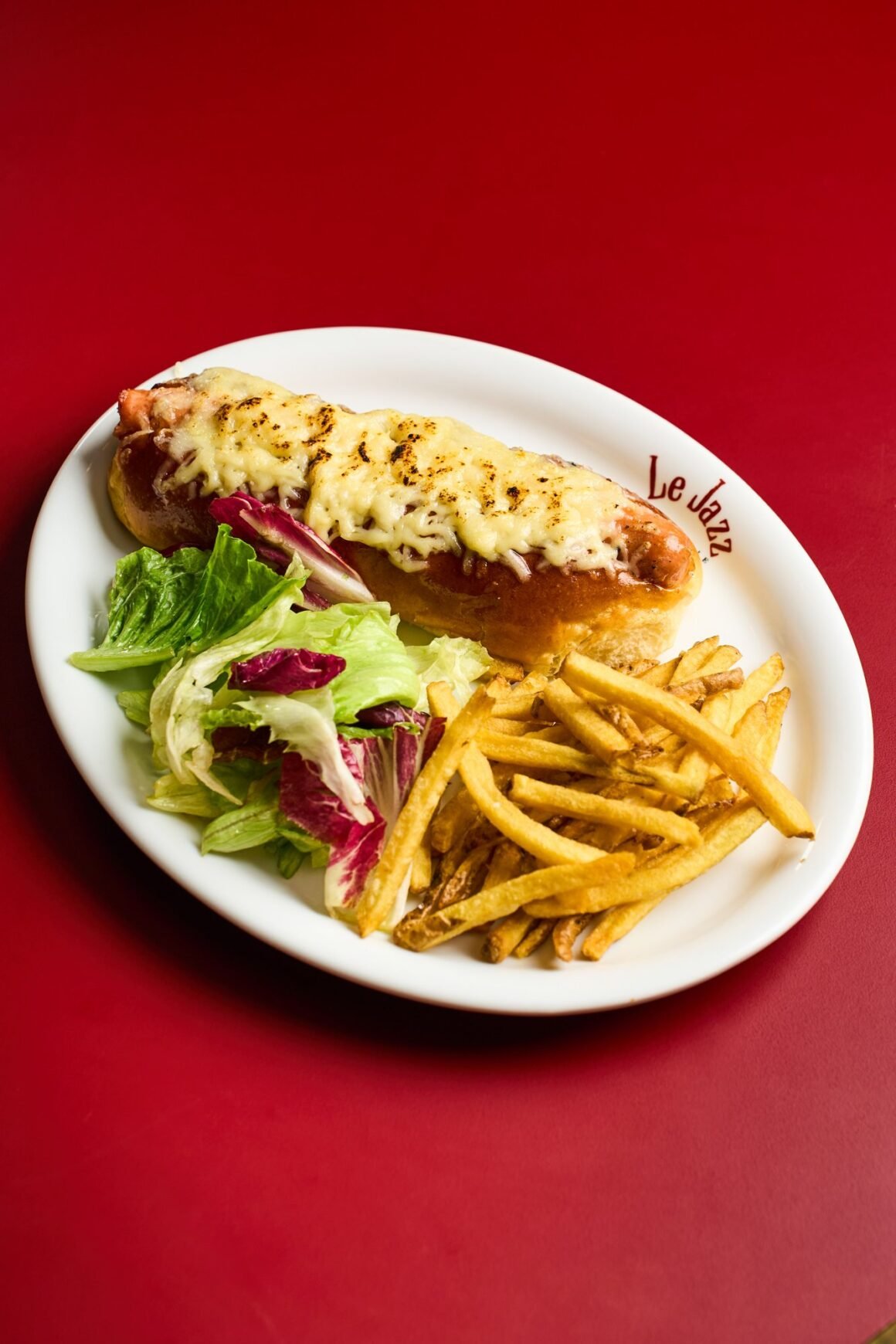 Cachorro-quente com salsicha gratinada com queijo gruyère no pão brioche, servido com batatas fritas e salada verde em prato branco sobre mesa vermelha