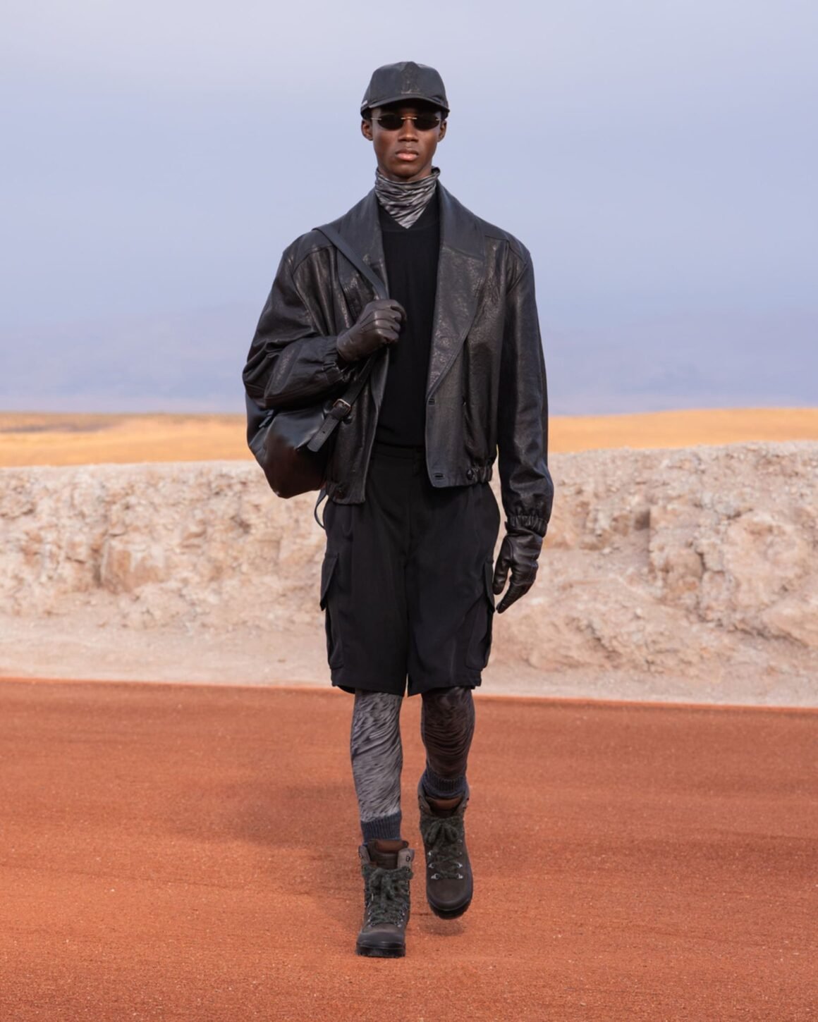 Modelo desfila com jaqueta de couro preta, bermuda cargo, luvas e botas no deserto de Agafay, na coleção Dreams of the Dune