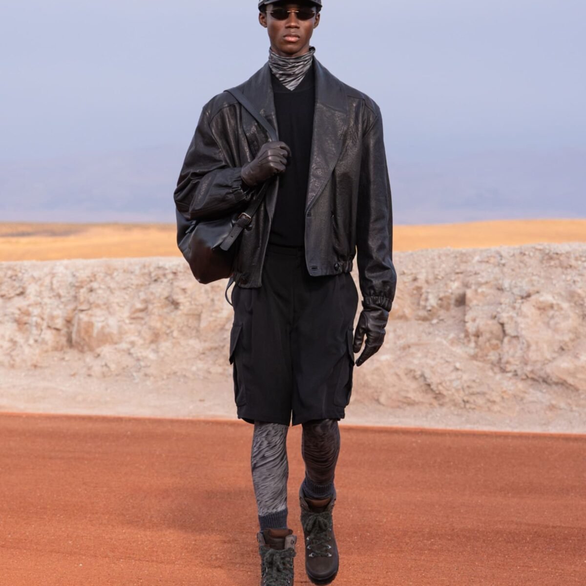 Modelo desfila com jaqueta de couro preta, bermuda cargo, luvas e botas no deserto de Agafay, na coleção Dreams of the Dune