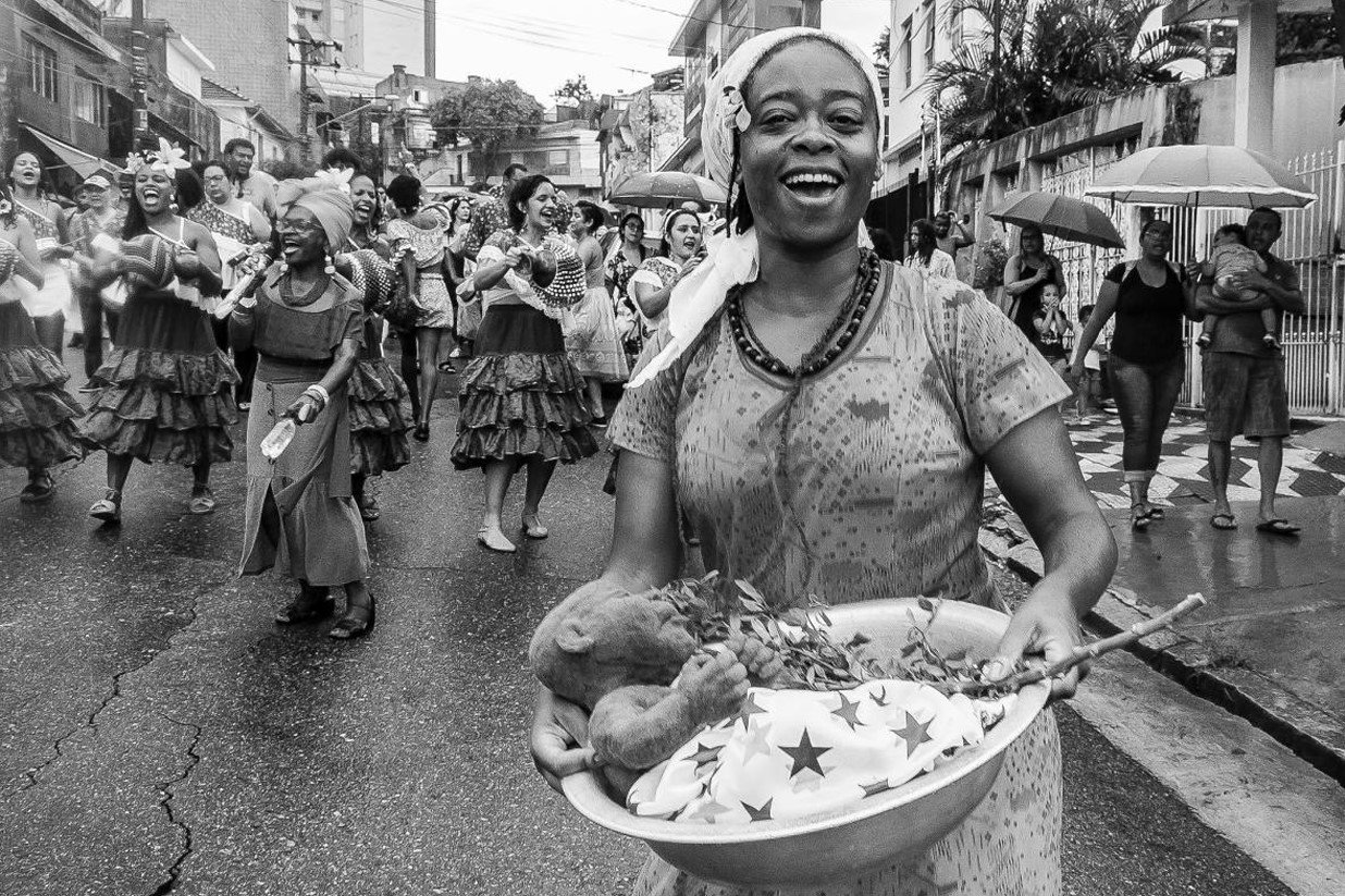 Fotografia em preto e branco de um cortejo de rua, com uma mulher sorridente à frente carregando uma bacia e outras mulheres de saias rodadas dançando atrás