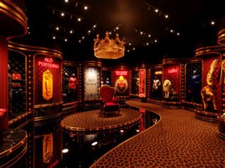 Projeção da exposição Sentimento Louco, com corredor de veludo vermelho, letreiros em neon com títulos de músicas, figurinos em vitrines e uma coroa dourada suspensa no centro