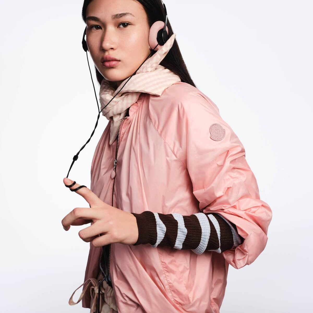 Modelo com jaqueta corta-vento rosa da Moncler, lenço listrado e fones de ouvido com fio