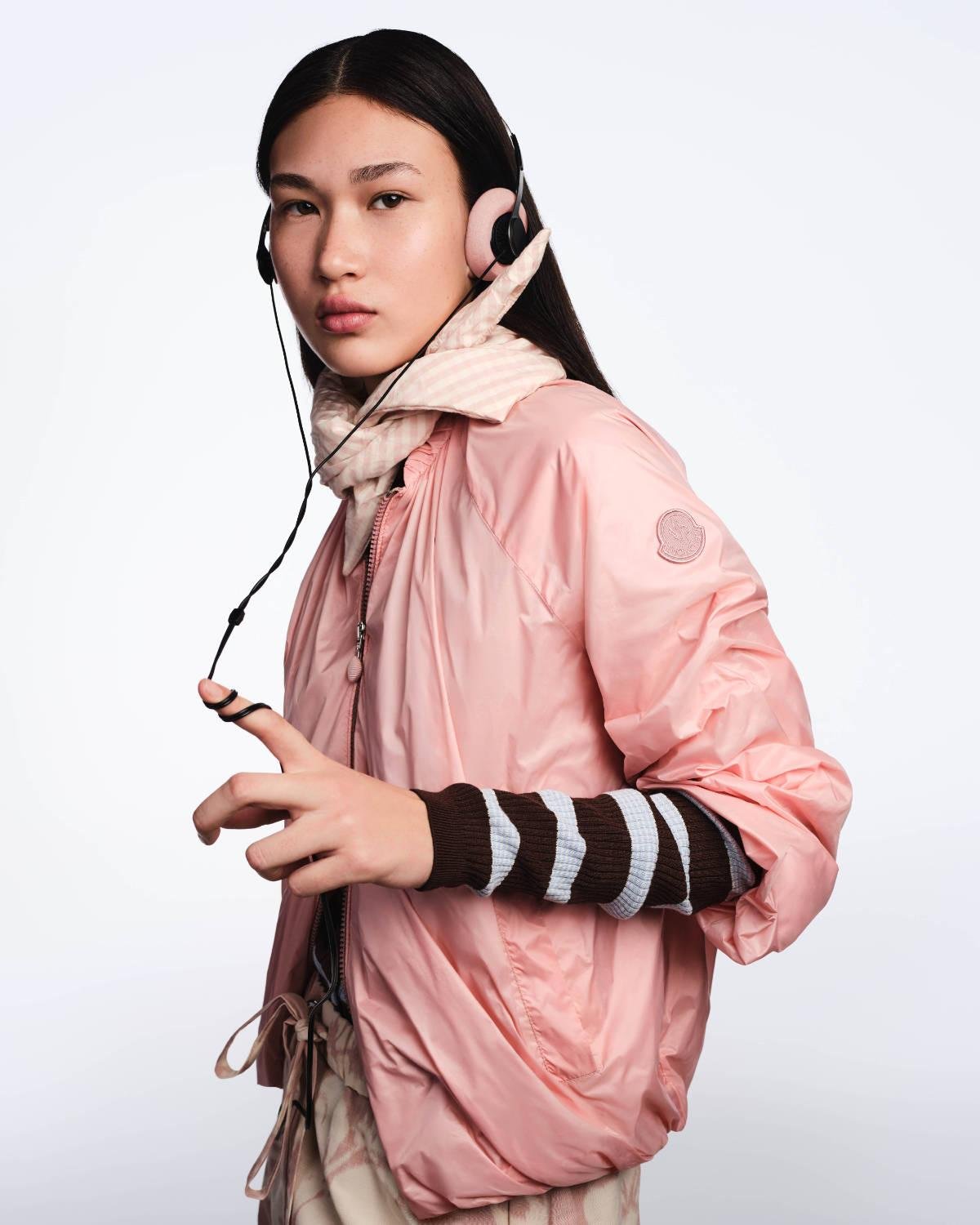 Modelo com jaqueta corta-vento rosa da Moncler, lenço listrado e fones de ouvido com fio