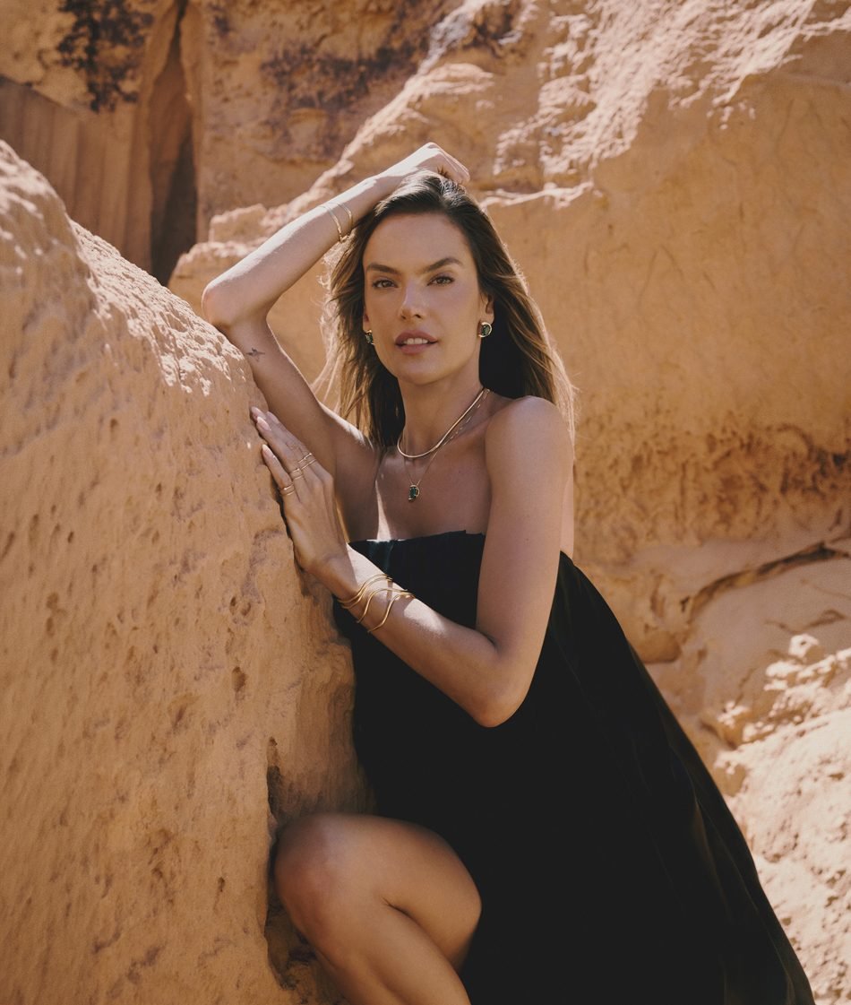 Alessandra Ambrosio apoiada em rochas alaranjadas, usando brincos, colar e pulseiras douradas com pedras verdes da Morana