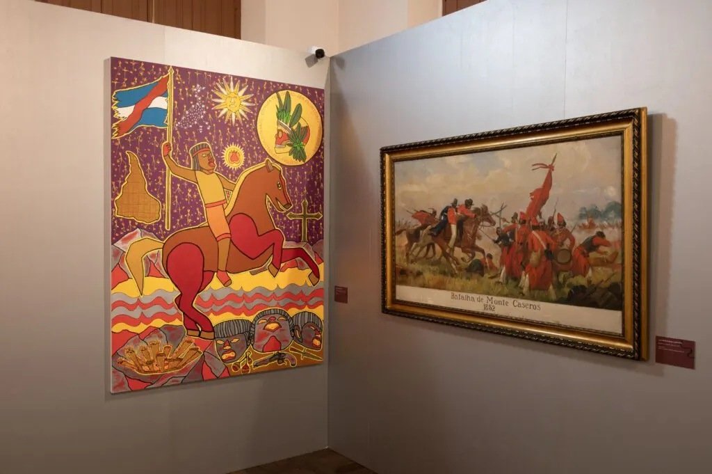 Sala da exposição com duas obras: à esquerda, pintura colorida de um guerreiro a cavalo segurando uma bandeira; à direita, tela emoldurada em dourado da Batalha de Monte Caseros, de 1852