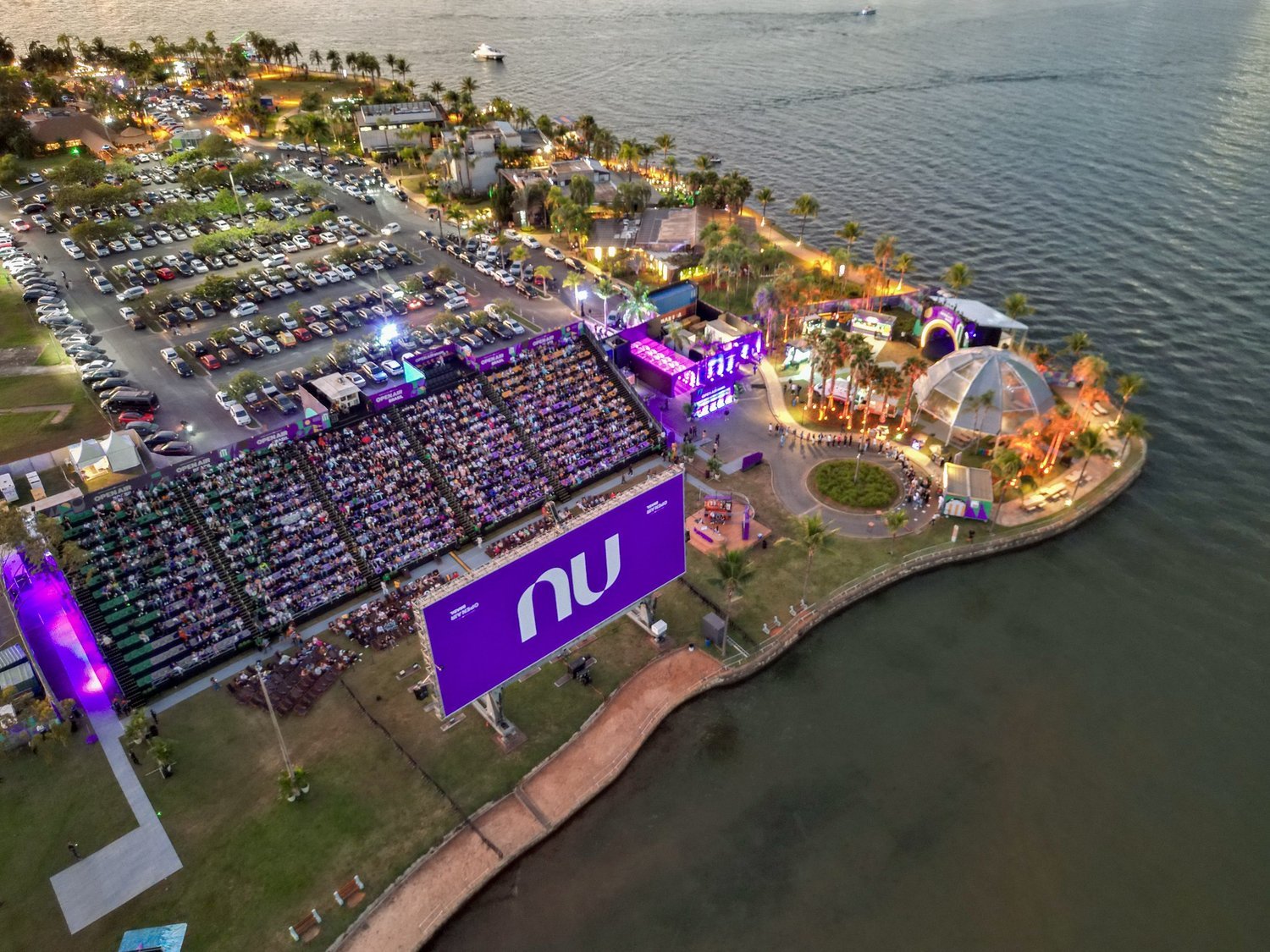 Vista aérea do Open Air Brasil ao entardecer, com a arquibancada diante da grande tela e a estrutura do evento à beira de um lago