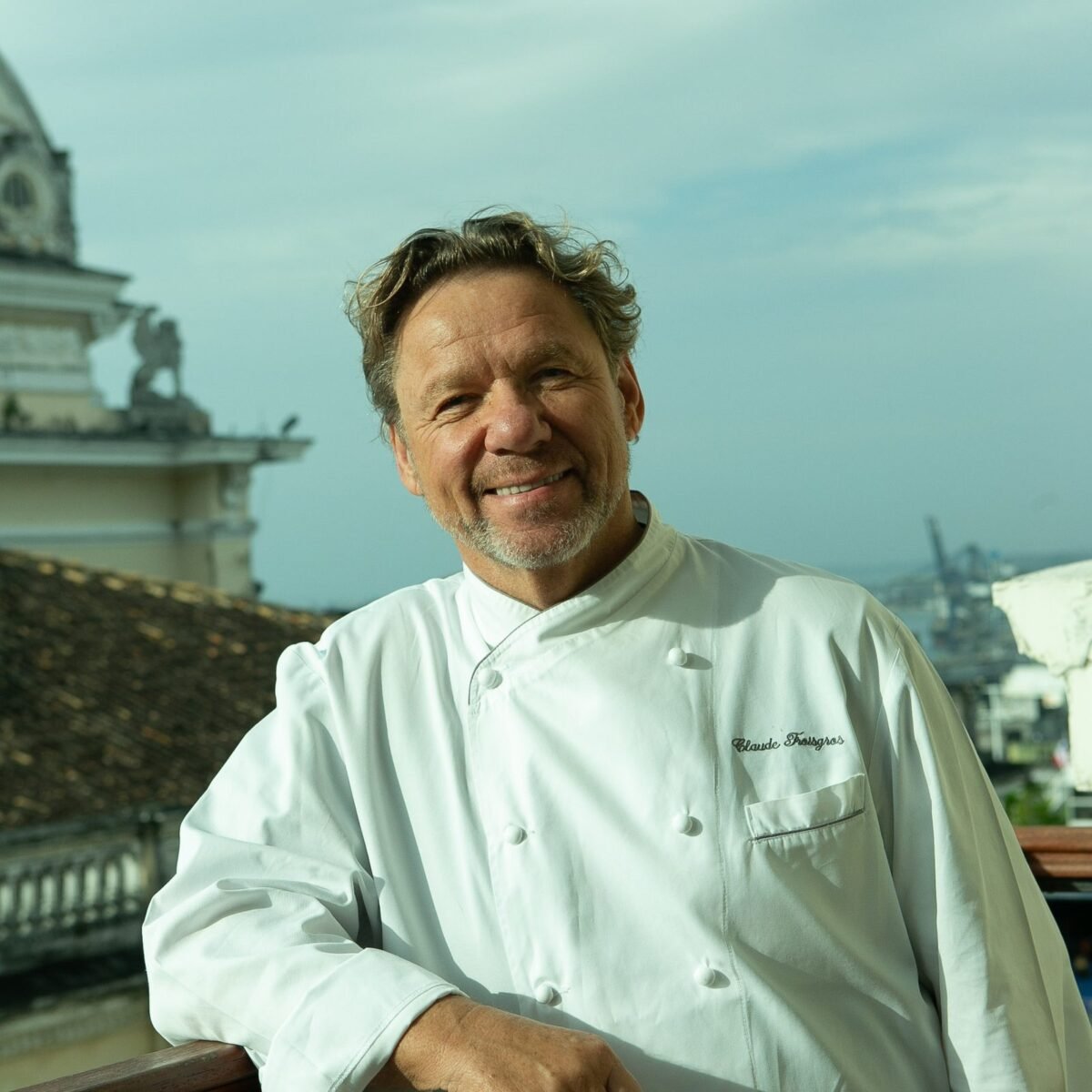 Retrato do chef Claude Troisgros, de dólmã branco, em varanda com vista para construções históricas de Salvador