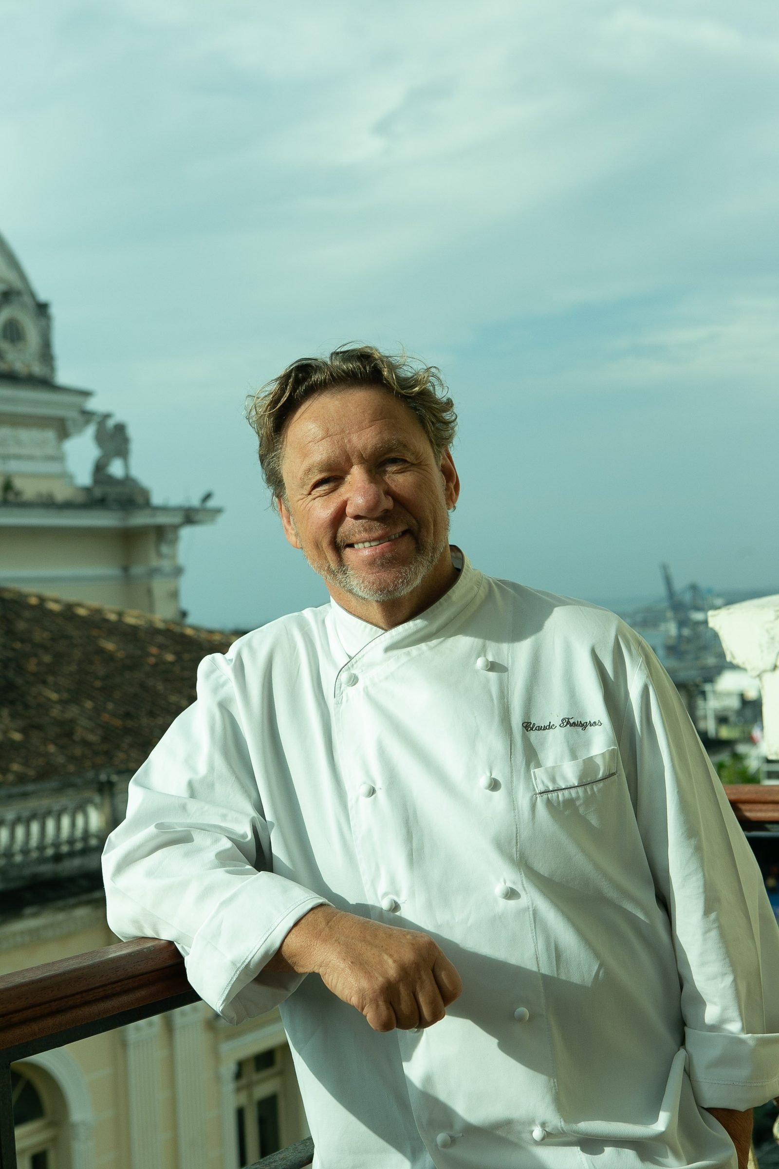 Retrato do chef Claude Troisgros, de dólmã branco, em varanda com vista para construções históricas de Salvador