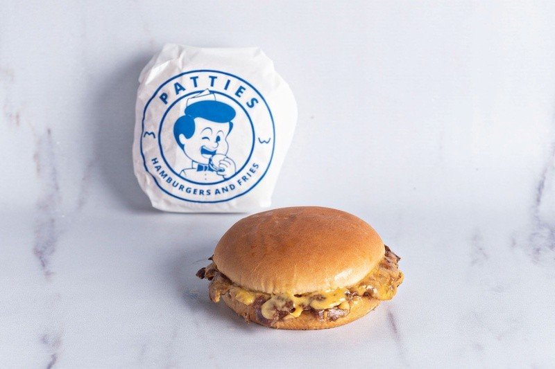 Cheeseburger com carne e queijo americano derretido em pão brioche, ao lado da embalagem de papel com a marca Patties, sobre bancada de mármore branco