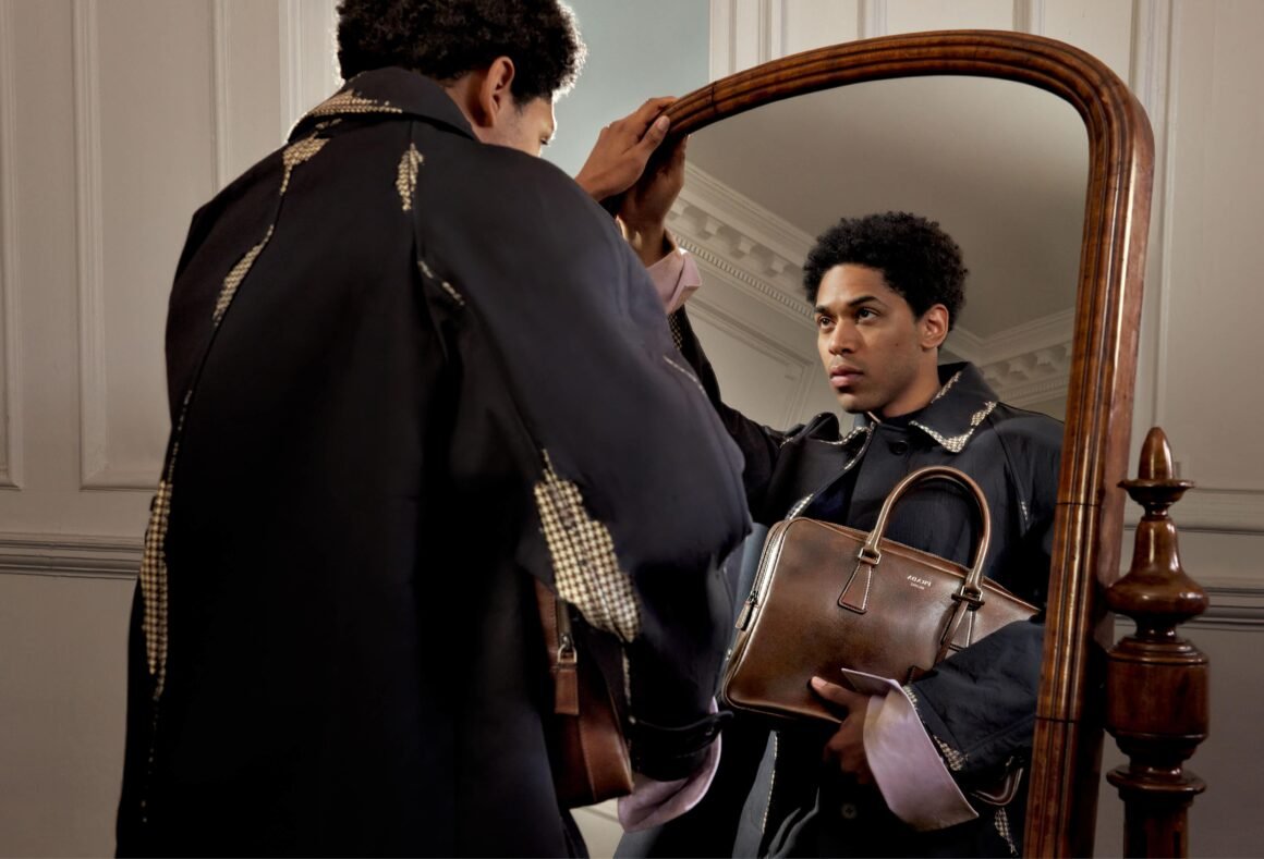 Modelo com sobretudo preto da Prada e bolsa de couro marrom diante de um espelho antigo.