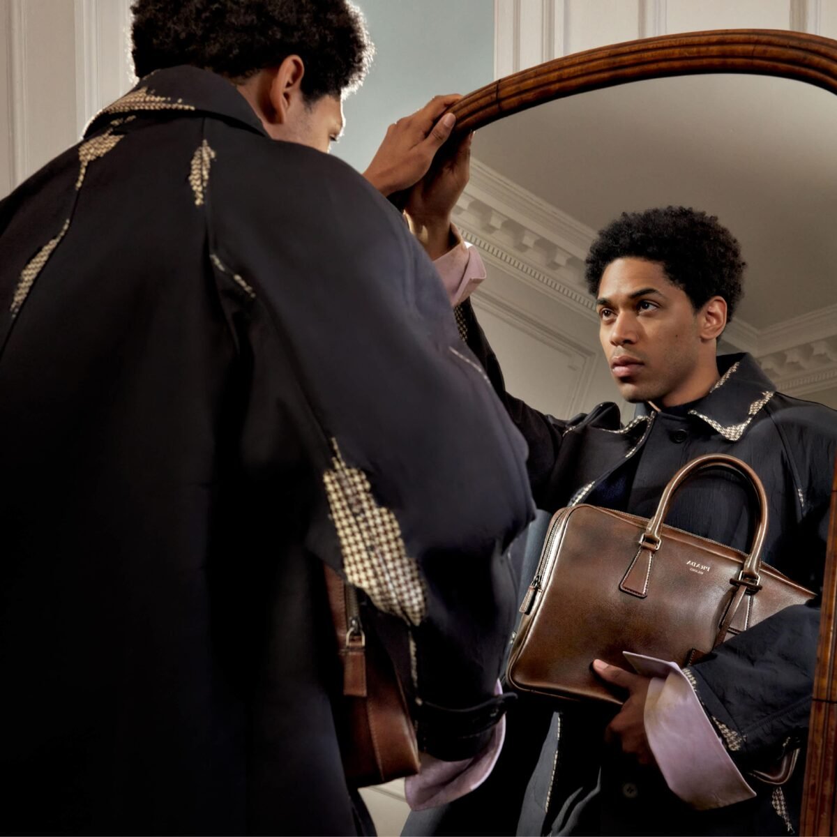 Modelo com sobretudo preto da Prada e bolsa de couro marrom diante de um espelho antigo.