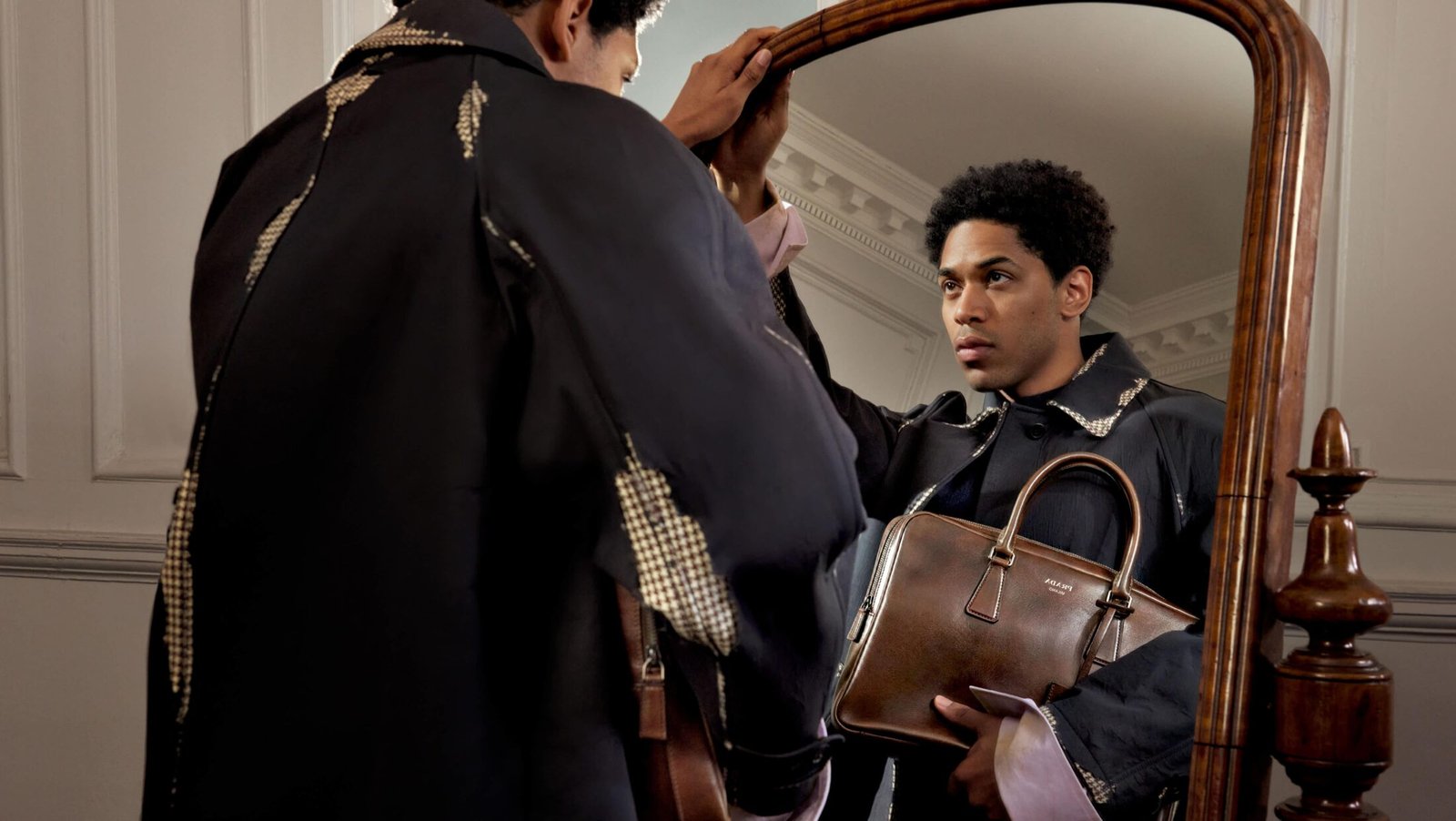 Modelo com sobretudo preto da Prada e bolsa de couro marrom diante de um espelho antigo.