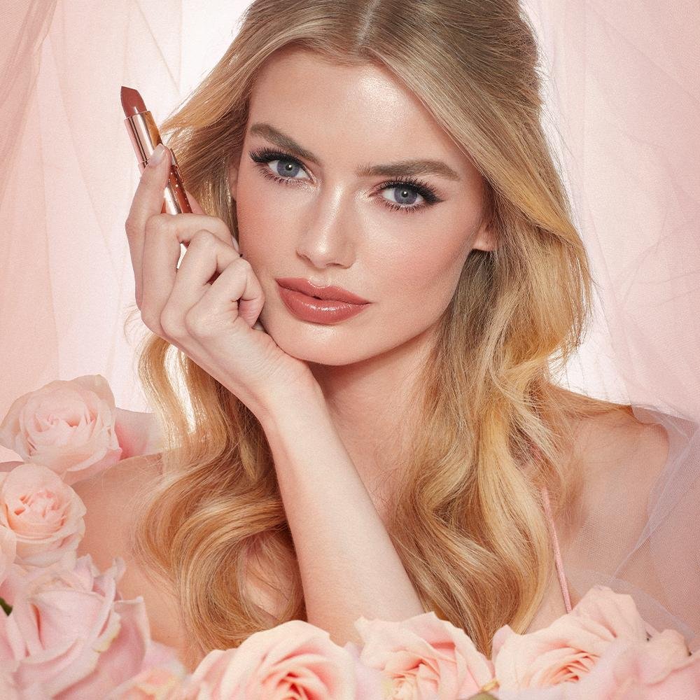 Modelo loira segura batom Charlotte Tilbury em tom nude, cercada por rosas cor-de-rosa