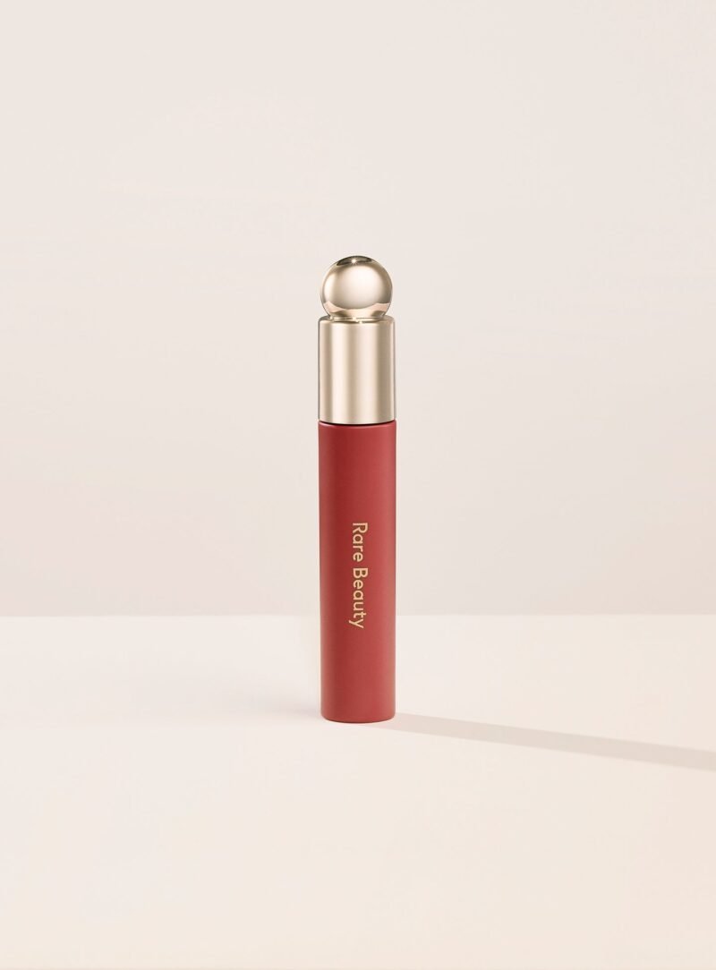 Frasco do Rare Beauty Soft Pinch Tinted Lip Oil com tampa dourada e óleo vermelho, de pé sobre fundo bege