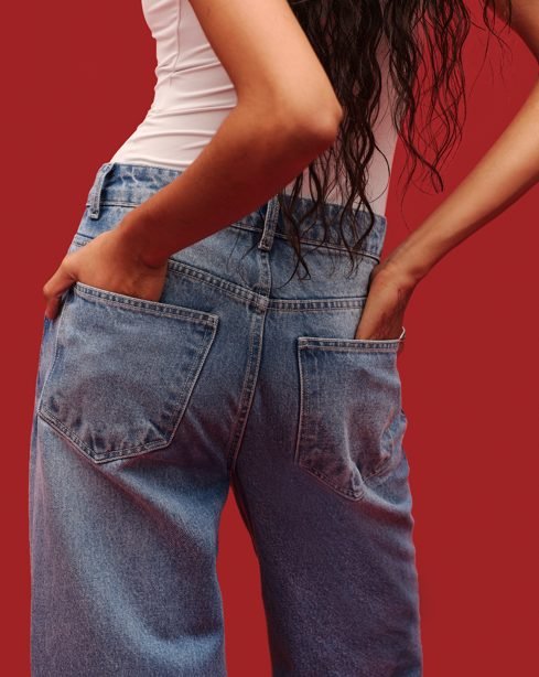 Detalhe de calça jeans wide leg da Renner em lavagem clara, vista por trás, com mãos nos bolsos e fundo vermelho