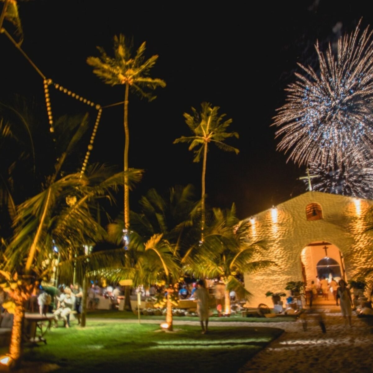 Capela iluminada entre coqueiros à noite, com fogos de artifício no céu e convidados vestidos de branco no gramado em frente