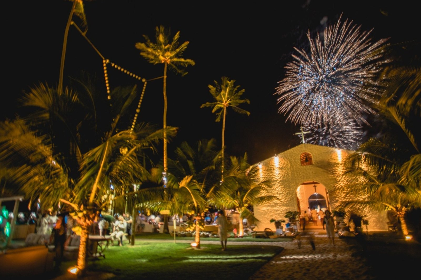 Capela iluminada entre coqueiros à noite, com fogos de artifício no céu e convidados vestidos de branco no gramado em frente