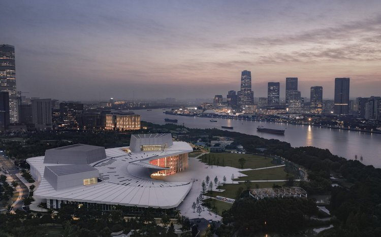 Vista aérea ao entardecer da Shanghai Grand Opera House, com cobertura espiralada branca às margens do rio Huangpu e o skyline de Xangai ao fundo
