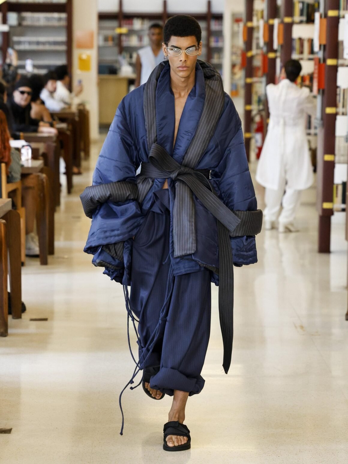 Modelo desfila com casaco acolchoado azul-marinho amarrado na cintura, da Handred, em uma biblioteca no SPFW N61