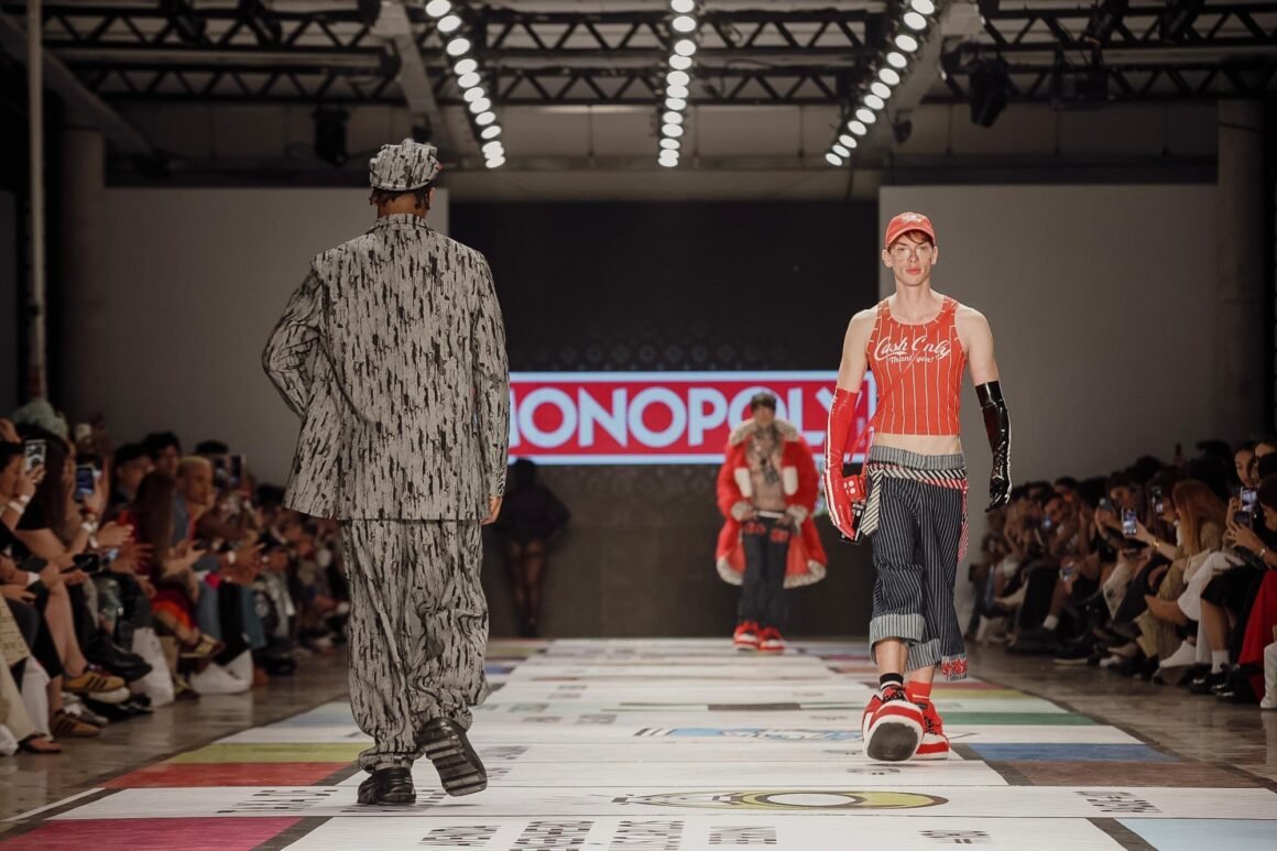 Modelos de Dario Mittmann na passarela com cenário de Monopoly, um de terno estampado e outro de regata vermelha e luvas, no SPFW N61