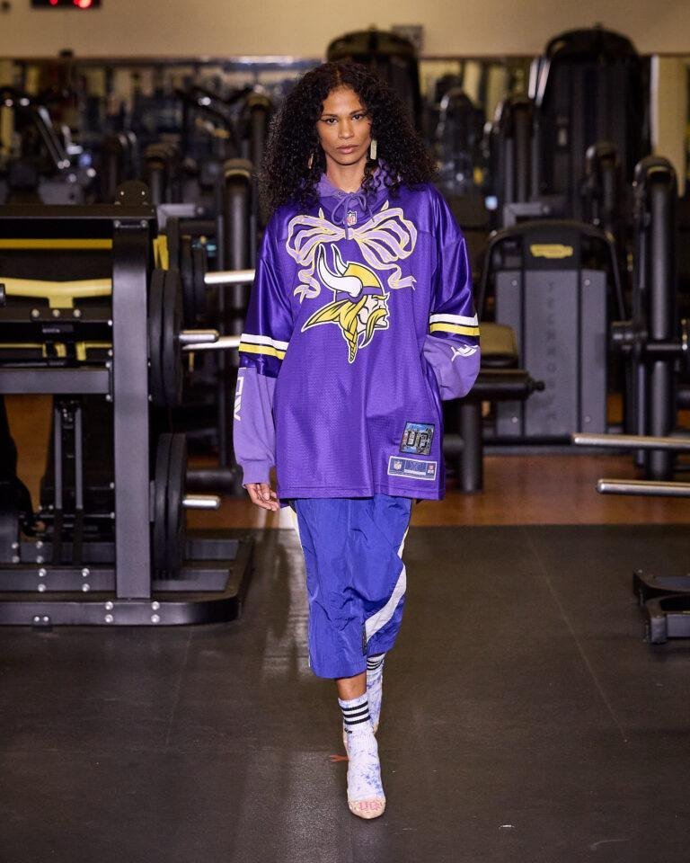 Modelo desfila com camisa de futebol americano roxa e calça azul da UÓ Upcycling em uma academia, no SPFW N61