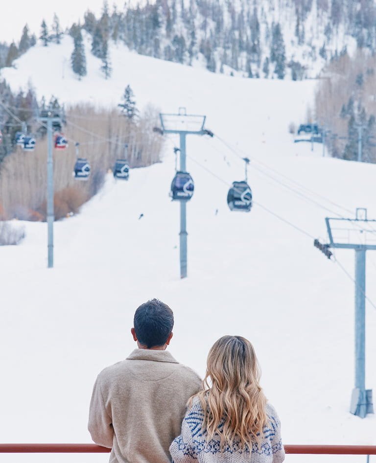 Casal de costas observa as gôndolas da Aspen Mountain cobertas de neve, em imagem de inverno do The Little Nell