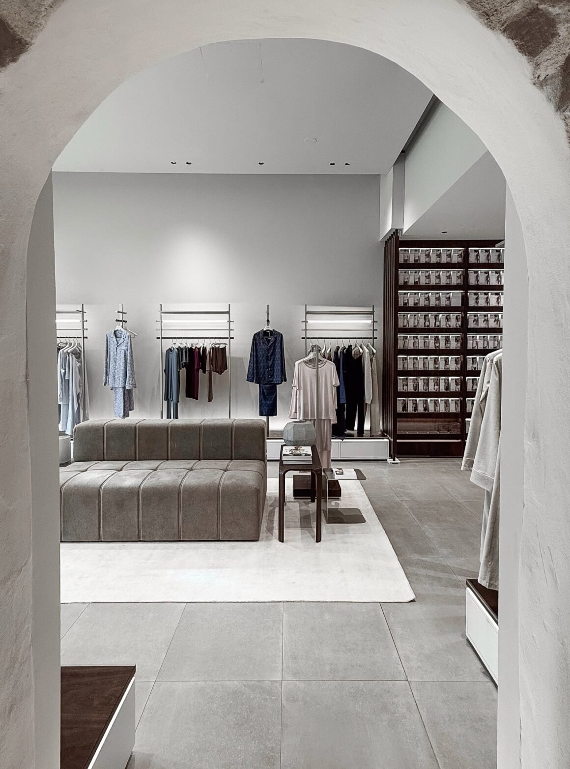 Interior da flagship Zimmerli em Zurique visto através de um arco, com araras de pijamas, sofá cinza e estante de madeira