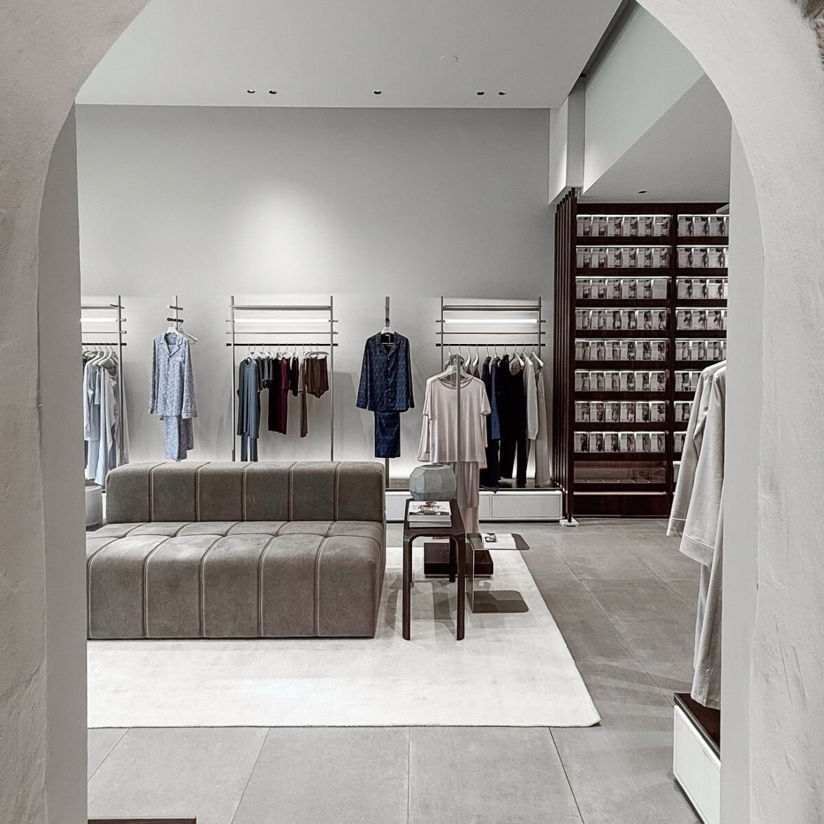 Interior da flagship Zimmerli em Zurique visto através de um arco, com araras de pijamas, sofá cinza e estante de madeira