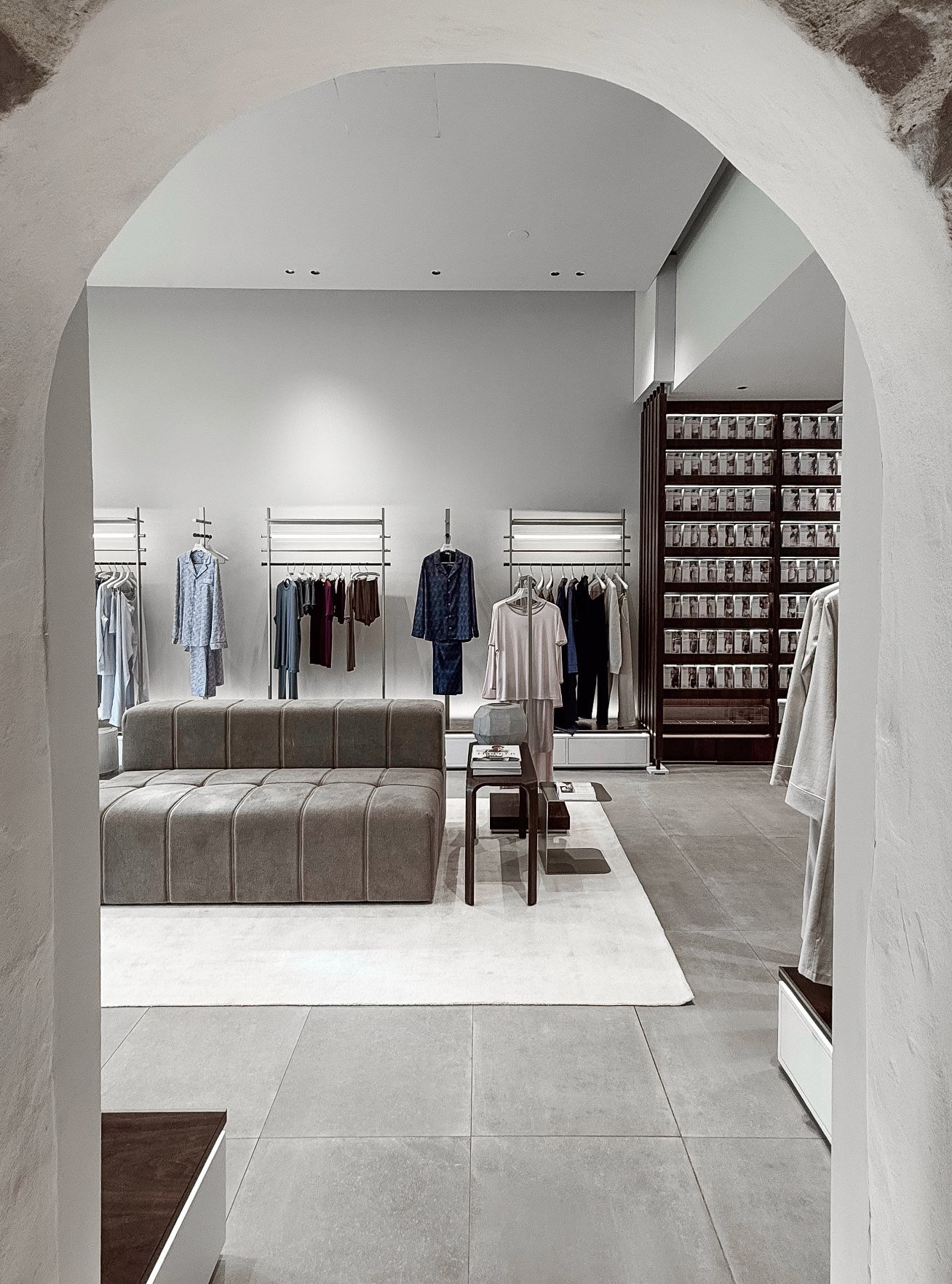 Interior da flagship Zimmerli em Zurique visto através de um arco, com araras de pijamas, sofá cinza e estante de madeira